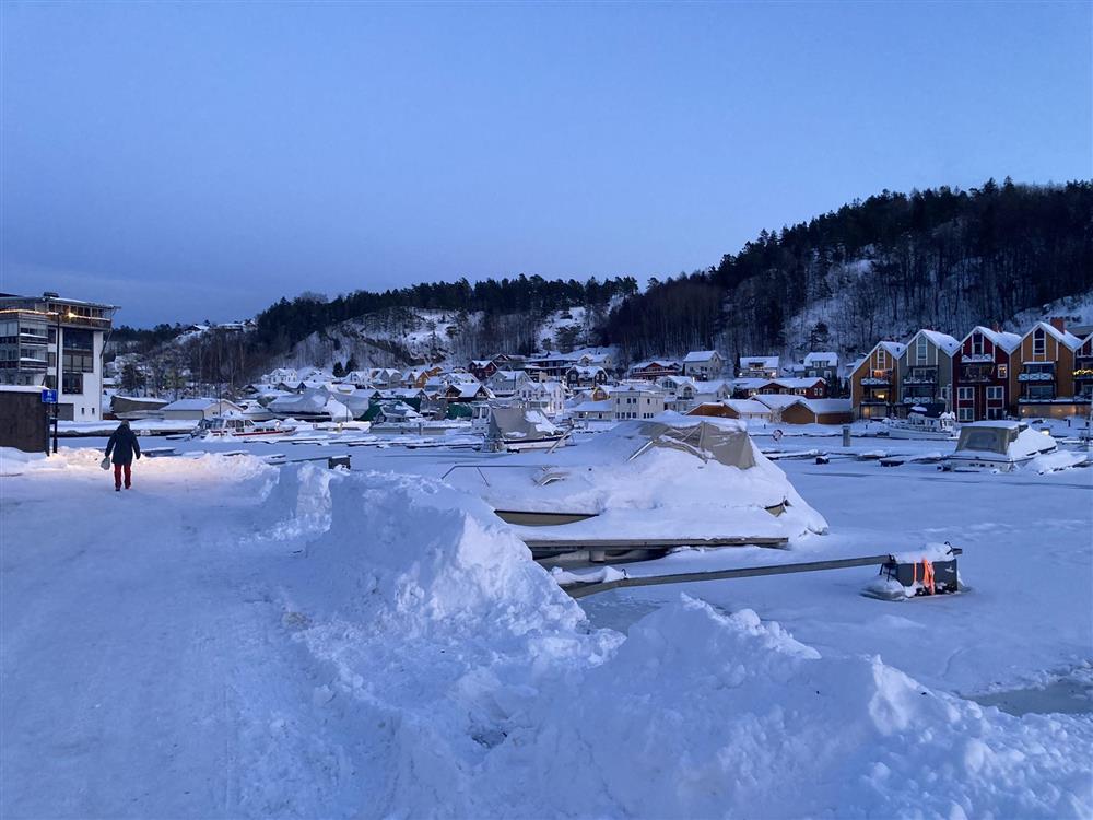 Person som går på måka gangvei ved sjø. Snøhauger og småbåtbrygger. Ås i bakgrunnen - Klikk for stort bilde