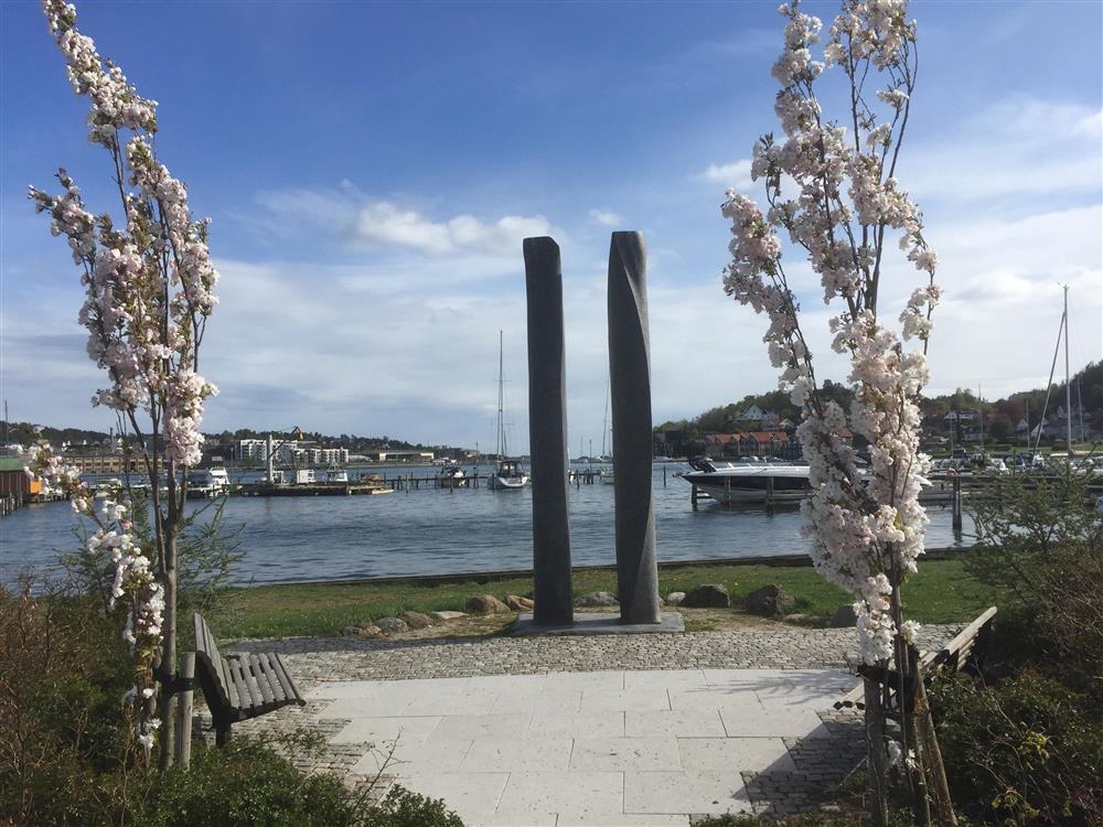 Sjø, blomstrende busker på hver side av skulptur der to søyler er reist - Klikk for stort bilde