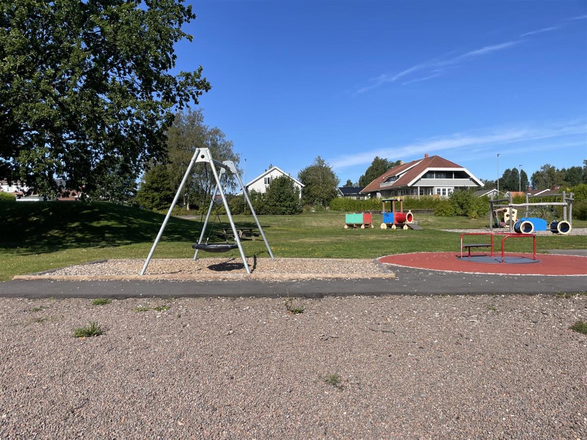 Lekeplass med huskestativ og karusell. Trær og andre lekeelementer i bakgrunn.  - Klikk for stort bilde