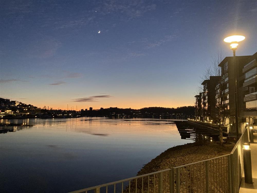 Speilblank sjø med bebyggelse på hver side. åser i bakgrunnen. Blå himmel og sjø og oransje rester av sol. Venus og nymåne på himmelen. - Klikk for stort bilde